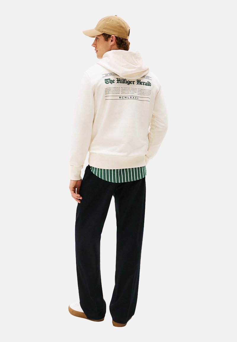 Hombre con una gorra beige, sudadera blanca con el texto "The Hilfiger Herald", camiseta a rayas verdes, pantalones negros y zapatillas blancas, de pie de espaldas.