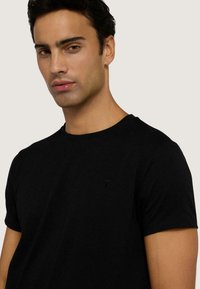 Czarna bawełniana koszulka z okrągłym dekoltem i krótkimi rękawami. Posiada małe, dyskretne logo na lewej piersi. Gładka tekstura.