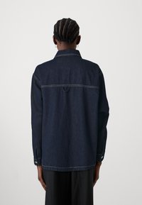 Mörk denimskjorta med rak passform, synliga sömnadsdetaljer, knäppte ärmslut och klassisk krage. Har en ögla bak.
