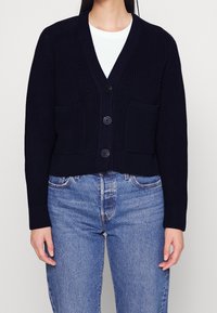 Marinblå kort cardigan i strukturerad stickad kvalitet, med djup V-ringning, tre knappar och två framfickor. Matchad med blå jeans.