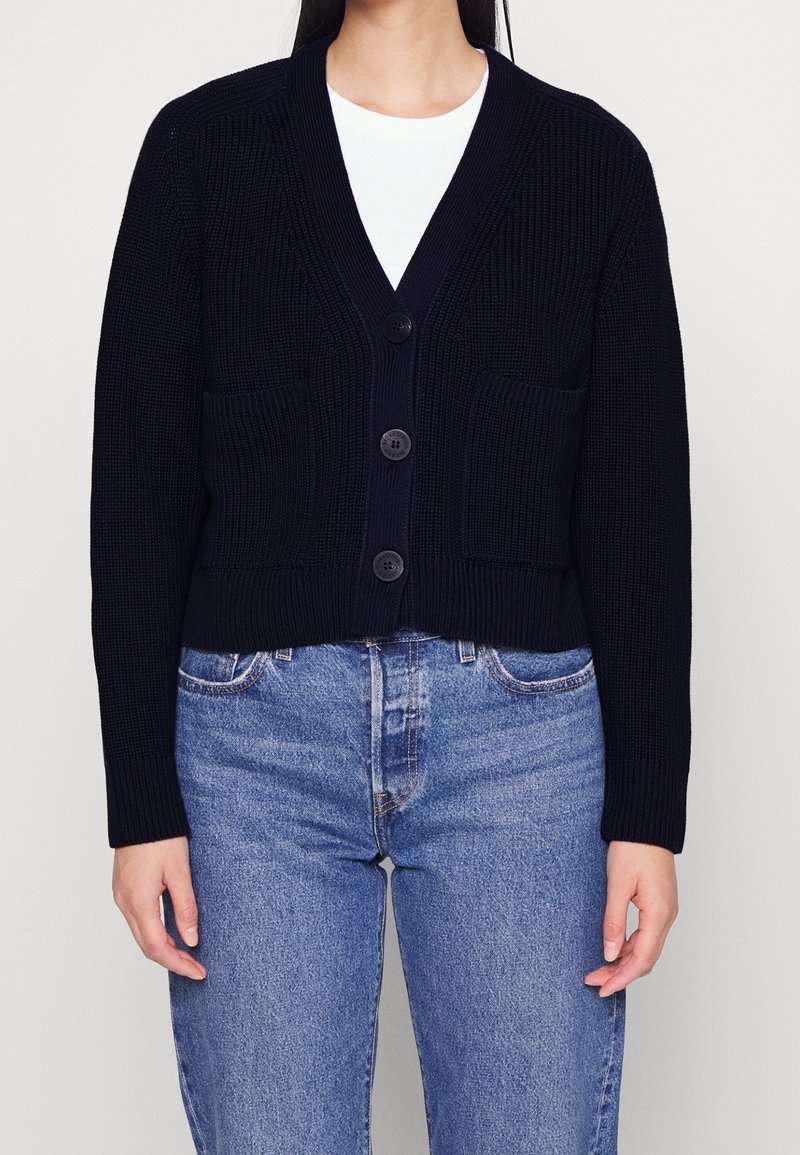 Marinblå kort cardigan i strukturerad stickad kvalitet, med djup V-ringning, tre knappar och två framfickor. Matchad med blå jeans.