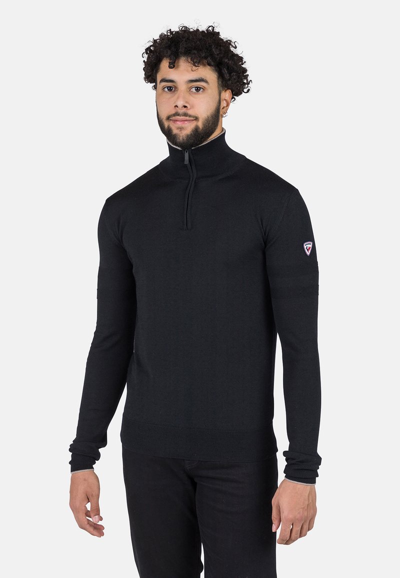 Maglione nero con zip al collo realizzato in tessuto a maglia, caratterizzato da maniche lunghe, design aderente e una piccola etichetta con logo sulla parte superiore del braccio.