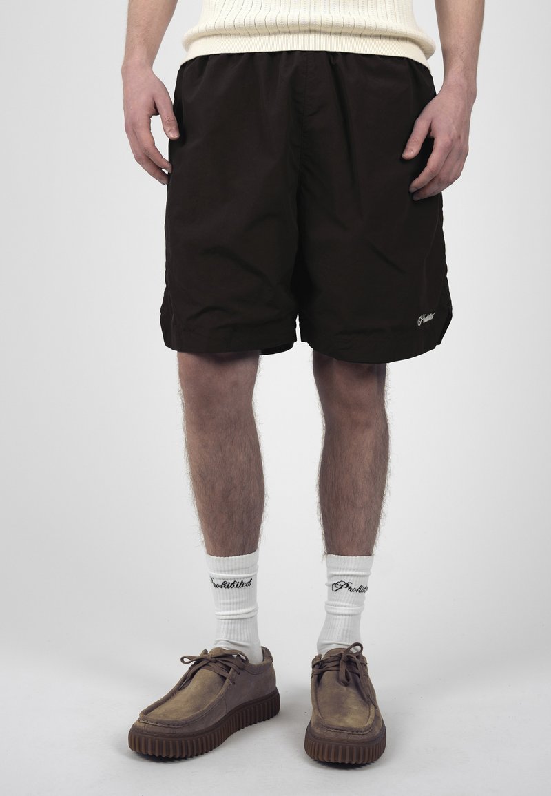 Personne portant un short noir, des chaussettes blanches avec du texte noir et des chaussures marron à lacets, debout devant un fond uni.