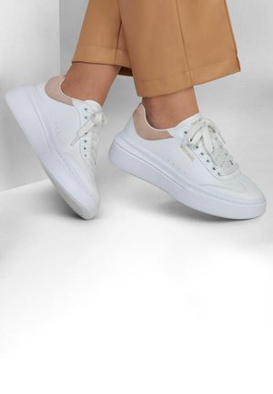 Baskets en cuir blanc avec des accents de talon beige portées avec un pantalon court beige contre un fond gris et blanc en angle.