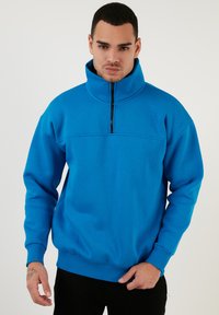 Sudadera azul de cuello alto, con cierre a media cremallera, fabricada en un tejido texturizado y con puños y dobladillo acanalados. Presenta un diseño de color sólido.