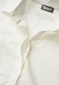 Camicia bianca con bottone in tessuto morbido, caratterizzata da un colletto classico, chiusura con bottone superiore e una leggera texture. Etichetta del marchio visibile all'interno.