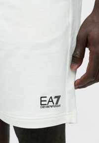 Tuvplānā tumšas ādas roka, kas pieskaras baltu EA7 Emporio Armani šortu apakšmalai.