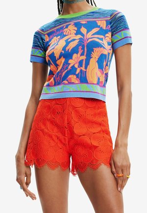Kvinde iført en blå og lilla blomstret kortærmet top og lyse orange blondershorts med store blomstermønstre.