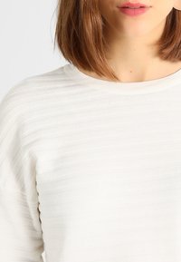 Witte textuur sweatshirt met horizontaal geribbelde ontwerpen en een ronde halslijn. Close-up focus op de stofdetails en bovenlichaam.