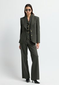 Costume taillé vert olive avec une veste à double boutonnage, boutons dorés, et pantalon large, associé à des escarpins noirs à talons pointus et des lunettes de soleil.