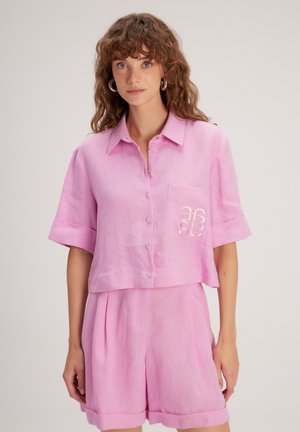 Camicia button-up in lino rosa con design cropped, maniche corte e una tasca a toppa con dettagli ricamati. Abbinata a pantaloni corto coordinati.