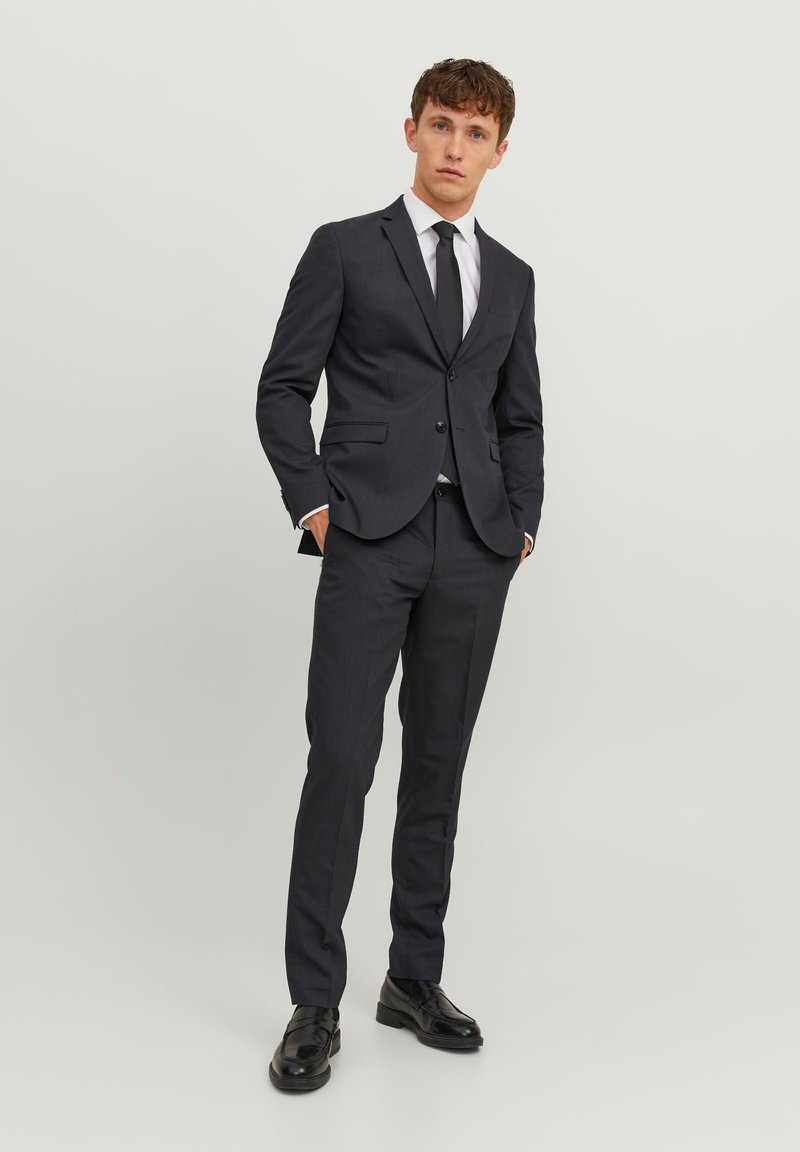 Completo grigio scuro con taglio slim, con rever a punta, due bottoni e pantaloni coordinati. Indossato con una camicia bianca e una cravatta nera, scarpe lucide.