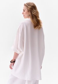 Luchtige, oversized witte blouse met opgerolde mouwen, gladde textuur en een licht gebogen zoom. Draag het met gelaagde armbanden om de pols.