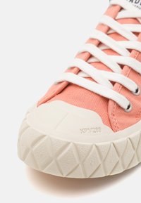 Koralowe sneakersy na płaskiej podeszwie z białymi sznurowadłami, okrągłymi oczkami i teksturowaną gumową podeszwą o geometrycznym wzorze. Wyraźne logo na języku.