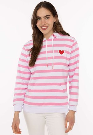 Junge Frau lächelt, trägt einen rosa-weiß gestreiften Hoodie mit einem roten Herz auf der Brust, kombiniert mit weißen Hosen.