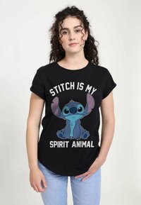 T-shirt en coton noir avec un graphisme d'un personnage de dessin animé bleu aux grandes oreilles, texte "STITCH EST MON ANIMAL SPIRITUEL" en blanc et argent.