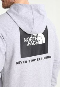 Hombre vestido con sudadera con capucha de color gris claro, que tiene un gran logotipo en negro y blanco de "The North Face" y el texto "Never Stop Exploring" en la parte posterior.