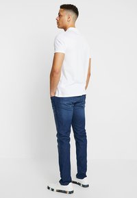 Witte polo met korte mouwen, gecombineerd met donkerblauwe spijkerbroek en witte sneakers met een gestipt zool. Van achteren gezien.