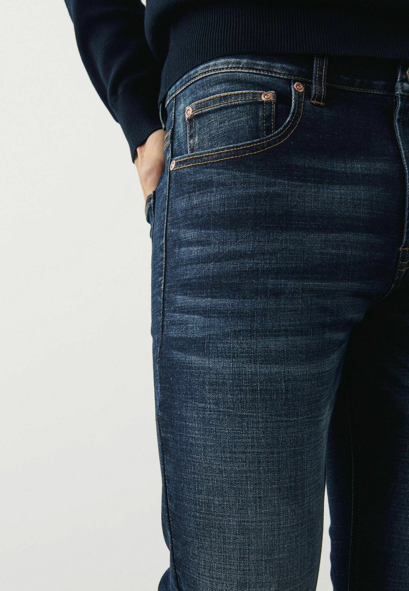 Jeans din denim albastru închis, cu o croială slim, detalii fine de uzură, și cusături portocalii contrastante, dotate cu buzunare standard și nituri metalice.