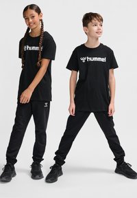 Doi copii poartă tricouri și pantaloni de trening negri Hummel, asortați. Tricourile au un logo alb; ambele sunt completate cu pantofi sport negri.