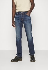 Jeans in denim blu scuro con vestibilità dritta, leggero sbiadimento sulle cosce e cuciture a contrasto. Abbinati a scarpe nere e una maglietta beige.