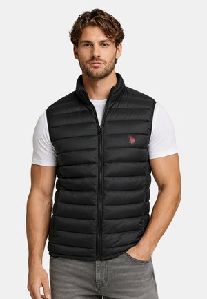 Man wearing black quilted vest over white short-sleeve shirt with hand in gray jeans pocket, neutral background.