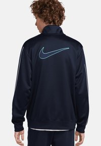 Chaqueta de pista Nike en color navy con un alto cuello, que presenta un logo color teal en la parte posterior y rayas laterales blancas. Textura suave, diseño atlético.