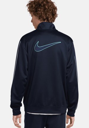Chaqueta de pista Nike en color navy con un alto cuello, que presenta un logo color teal en la parte posterior y rayas laterales blancas. Textura suave, diseño atlético.