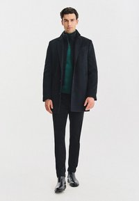 Czarny płaszcz o strukturalnym kroju, noszony na zielonym golfie, w połączeniu z czarnymi spodniami i botkami za kostkę, na jasnym tle.