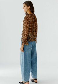 Blouse met luipaardprint, met ruches, doorzichtig materiaal en lange mouwen, gecombineerd met hoogatsinni lichte blauwe wijde jeans en zwarte slides.