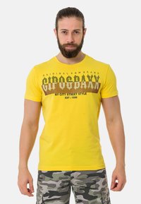 Geel katoenen T-shirt met korte mouwen, voorzien van gedrukt tekst in donkerbruin en zwart, gecombineerd met shorts in camouflagepatroon.