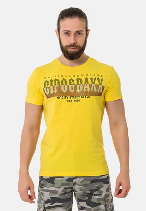 Gelbes Baumwoll-T-Shirt mit kurzen Ärmeln, das bedruckten Text in Dunkelbraun und Schwarz aufweist, kombiniert mit camouflagemusternden Shorts.