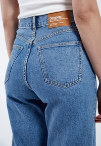 Ljust blå denimjeans med hög midja, synliga bakfickor och en brun läderlapp på baksidan, med orange sömnadsdetaljer.