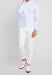 Camisa de botón azul claro de manga larga, pantalones blancos y zapatillas blancas con suelas de goma. La camisa presenta un pequeño logo en el pecho.