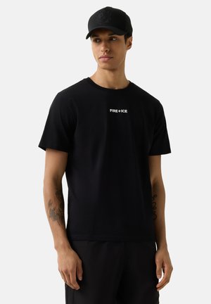 Ung mand iført en sort kasket og sort T-shirt med teksten "FIRE + ICE", stående foran en ensartet lys baggrund og kigger mod venstre.