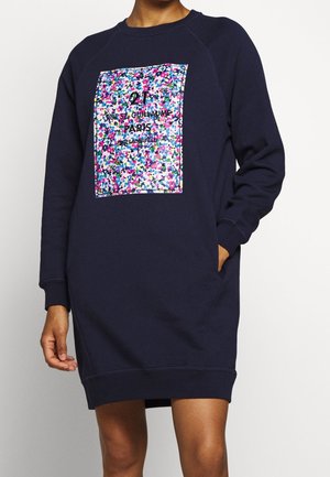 Marineblaues Sweatshirtkleid mit langen Ärmeln, buntem floralem rechteckigem Aufnäher und Paris-Schriftzug auf der Brust, Modell mit einer Hand in der Tasche.