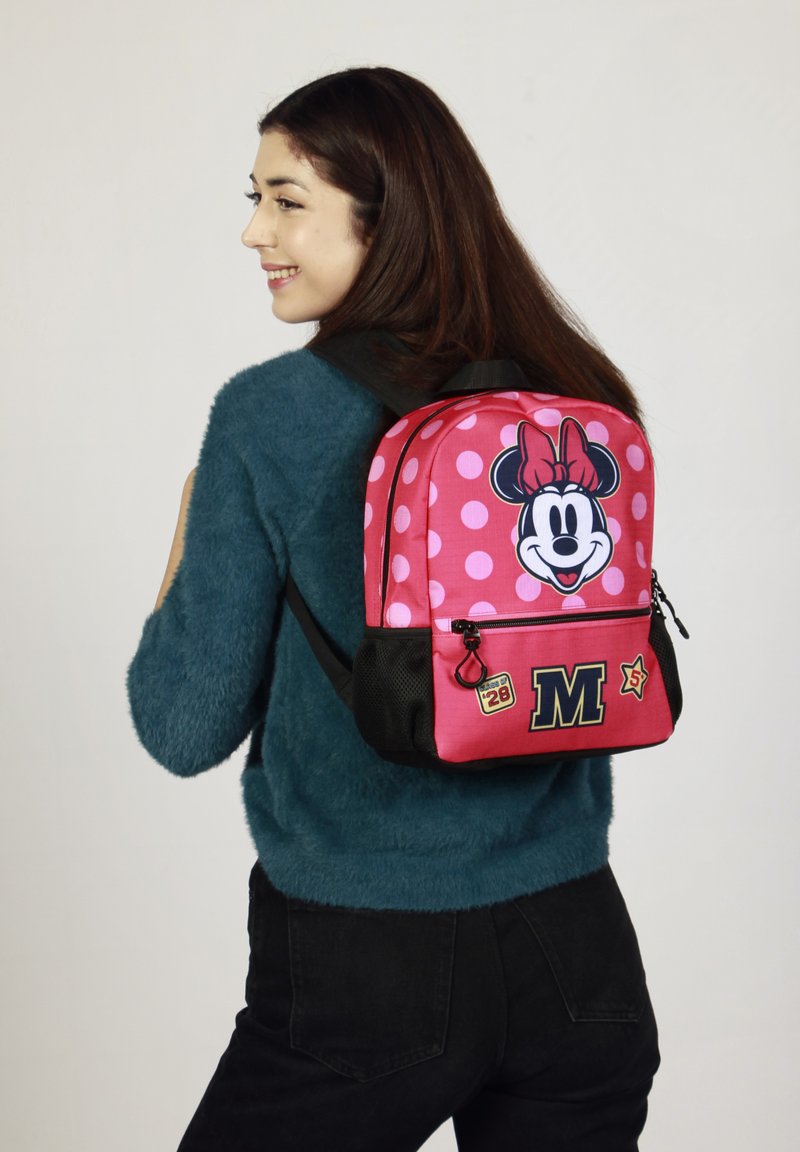 Mochila roja y rosa con lunares que presenta un gran gráfico de Minnie Mouse, acentos en negro y un bolsillo frontal con cremallera adornado con parches decorativos.