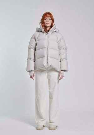 Donna con capelli rossi indossa una giacca a piumino grigio chiaro, pantaloni bianchi e scarpe beige, in piedi davanti a uno sfondo chiaro e uniforme.