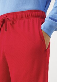 Personne portant un short de sport en maille rouge avec cordon de serrage, main dans la poche latérale, et une chemise à manches longues bleue avec manches retroussées.