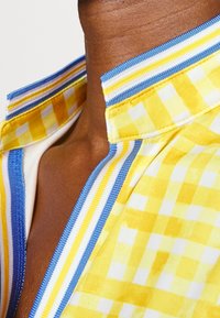 Detalj av en gul rutmönstrad skjorta med en blå och gul randig krage, som visar en slät textur och kontrasterande mönster.