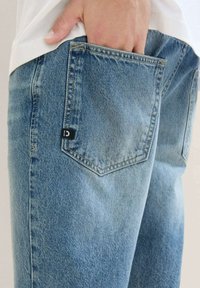 Vaaleansininen farkkutakki klassisella leikkauksella, jossa on takatasku ja musta label. Kankaassa on lievää tekstuurin vaihtelua.