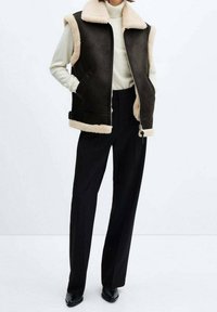 Veste marron sans manches avec un col et des bordures en fausse fourrure beige, fermeture éclair à l'avant, portée avec un pantalon et des chaussures noirs.