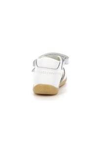 Sandalia de cuero blanca con acentos plateados, vista trasera. Presenta una correa ajustable y una suela de goma marrón claro, grabada con "Kickers".