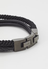 Le bracelet en cuir tressé noir présente un élégant fermoir en métal avec le logo "FOSSIL", alliant texture tressée et accents lisses.