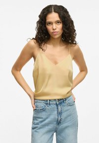 Camiseta de satén amarilla clara con escote en V y tirantes finos, combinada con jeans de cintura alta de color azul claro, con detalles desgastados.