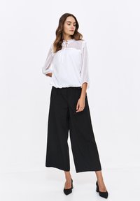 Biała bluzka w kropki z przezroczystymi, falbankowymi rękawami i wiązaniem przy szyi, w połączeniu z czarnymi, wysokimi spodniami o szerokich nogawkach i czarnymi czółenkami ze spiczastym noskiem.