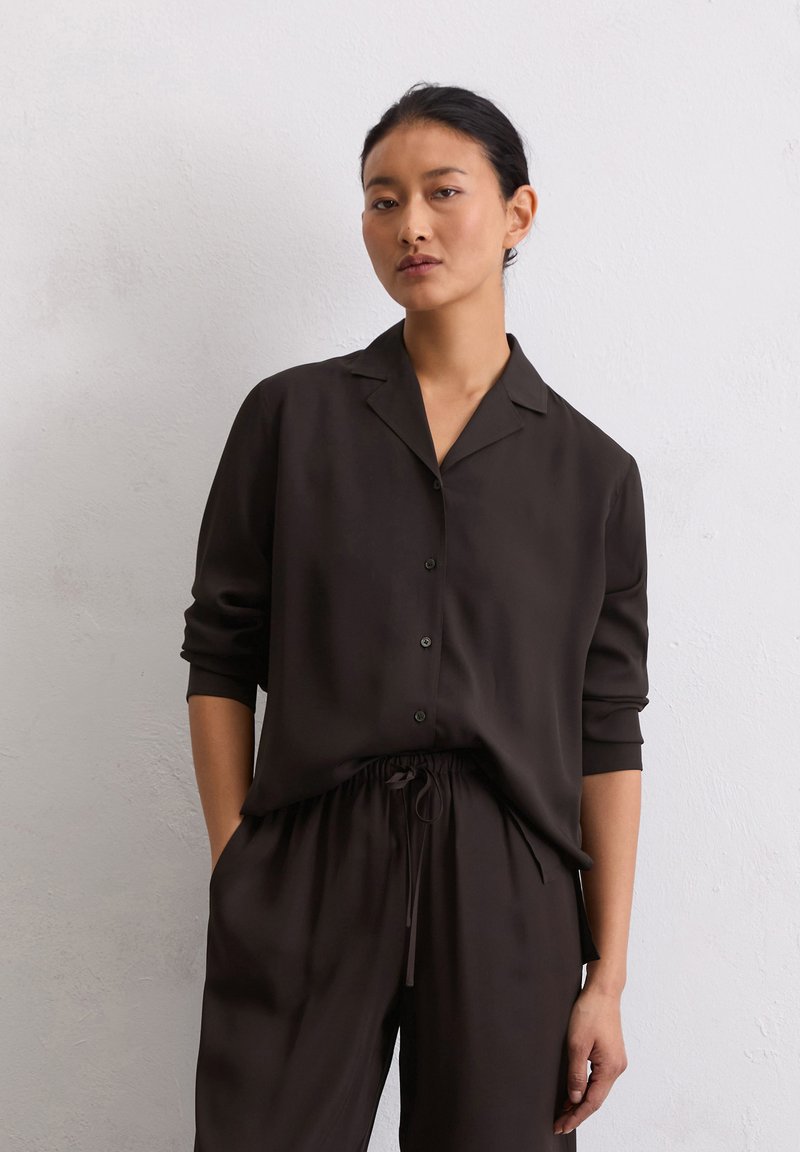 Camicia button-up di un marrone scuro setoso con colletto a contrasto e maniche arrotolate, abbinata a pantaloni con cordino dello stesso colore. Texture liscia in tutto.