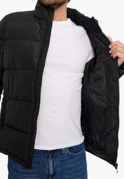 Giacca imbottita nera con zip frontale, colletto alto e maniche infilate, indossata sopra una camicia bianca e jeans blu. Texture liscia.