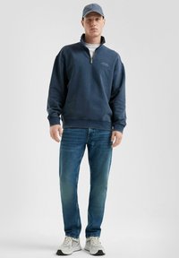 Marineblauer Reißverschluss-Hoodie mit gerippten Bündchen und Saum, kombiniert mit verwaschenen blauen Jeans und grauen Sneakers. Der Hoodie zeigt ein einfaches Logo.