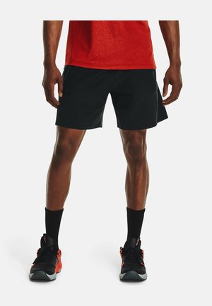 Homme debout, jambes écartées, portant un short de sport noir, des chaussettes noires, des chaussures de course rouges et noires, et un t-shirt rouge.